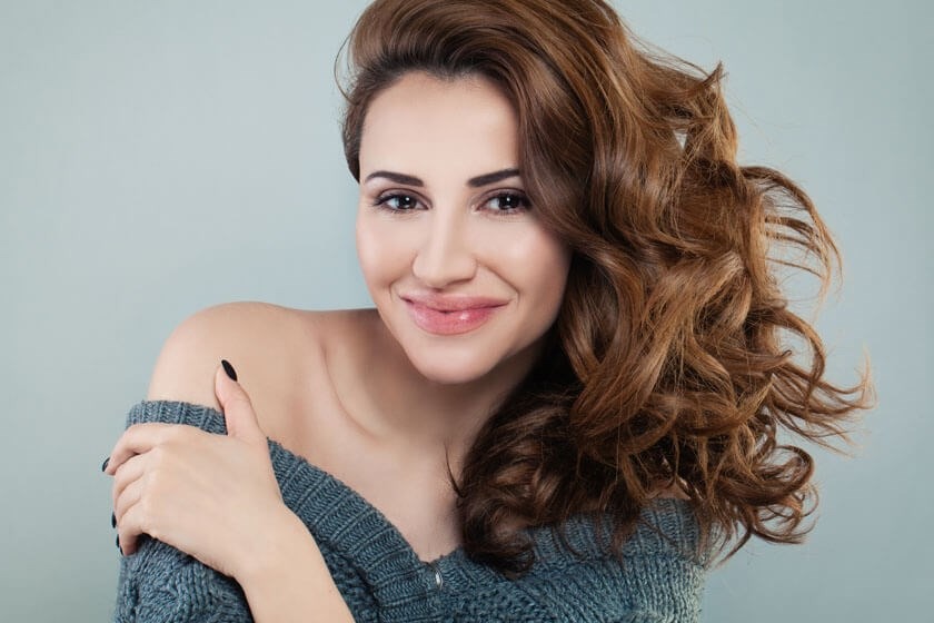 A woman with long, wavy brown hair and natural makeup smiles at the camera. She is wearing an off-the-shoulder blue sweater and is standing against a plain light gray background.