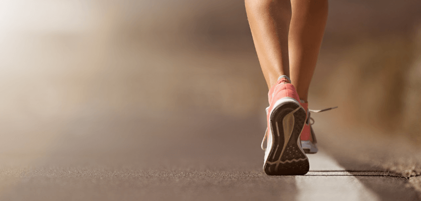 Close-up of a person walking or running on a road, wearing pink athletic shoes. Only their lower legs and feet are visible, with a blurred background.