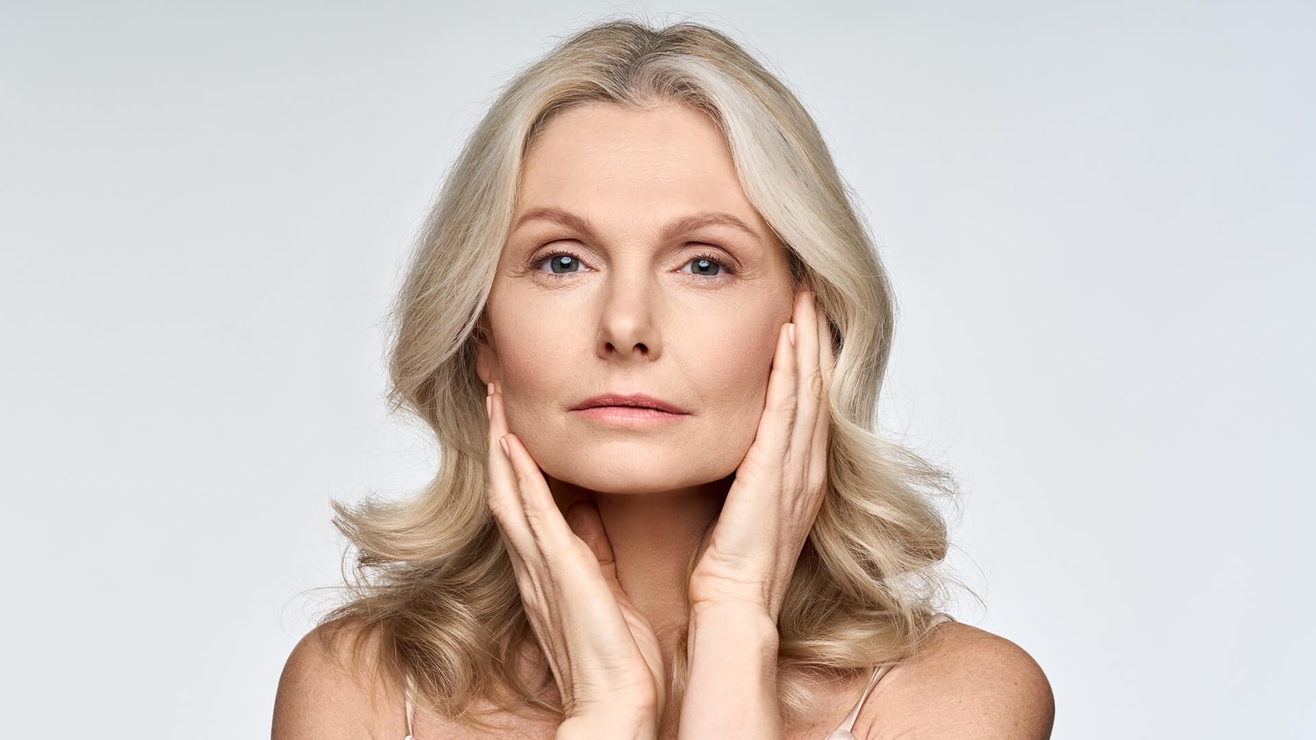 A mature woman with light blonde hair touches her face gently with both hands, looking directly at the camera with a calm expression against a plain, light background.