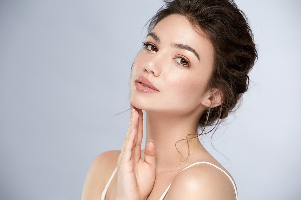 A young woman with light skin and dark hair in a loose updo poses against a light gray background, gently touching her chin with her hand. She has natural makeup and wears a white spaghetti strap top.