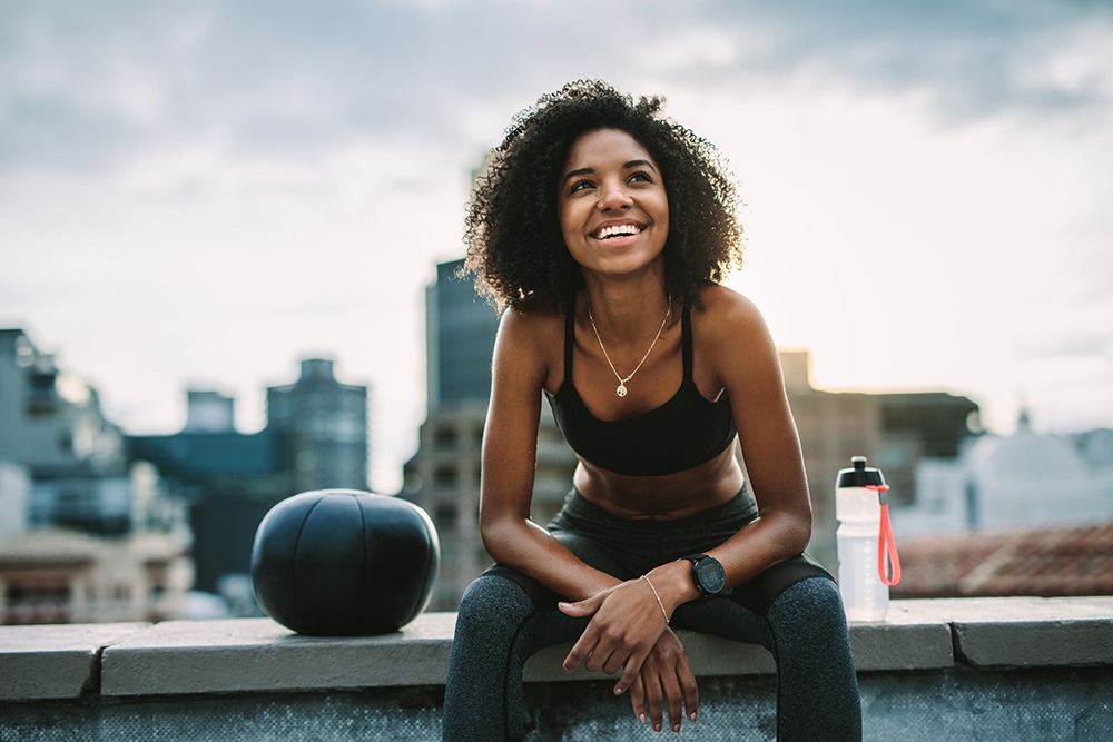 A woman in athletic wear sits on a rooftop ledge, smiling and looking up. She is next to a medicine ball and a water bottle, with a city skyline in the background.