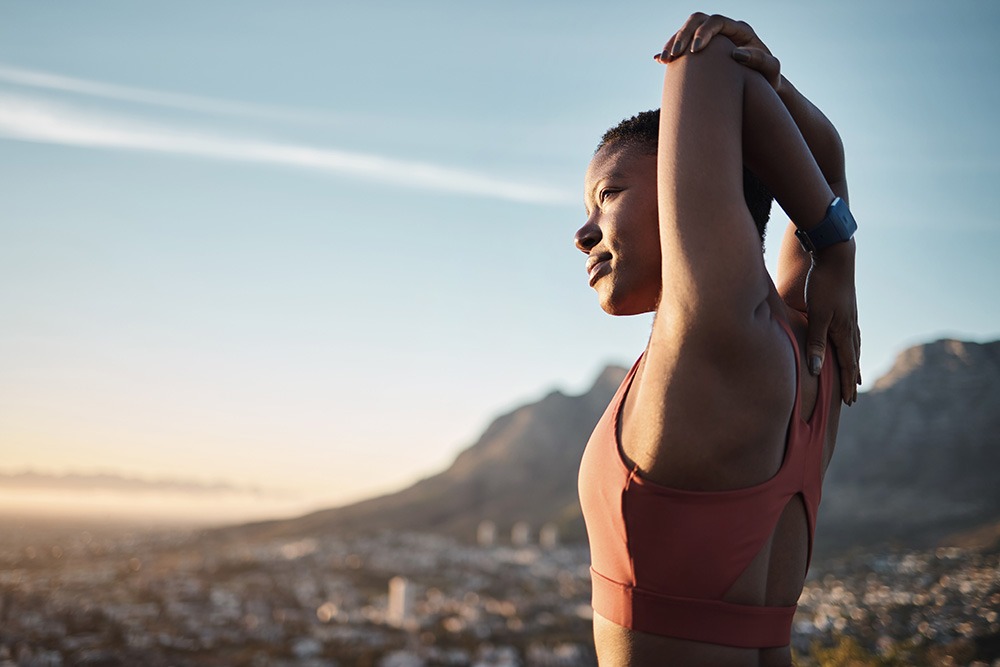 A woman in athletic wear stretches her arms overhead outdoors, with mountains and a cityscape visible in the background during sunrise or sunset.