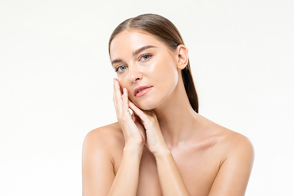 A woman with clear skin and straight brown hair poses against a white background, gently touching her face with both hands and looking at the camera. She has a natural makeup look and bare shoulders.