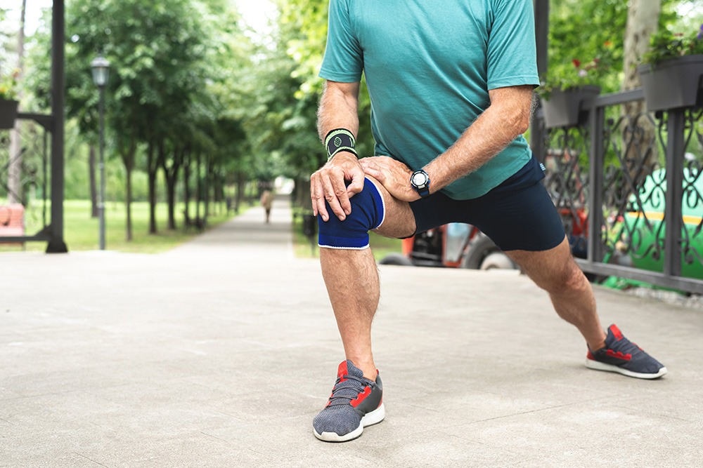 A person wearing a teal shirt, dark shorts, and sneakers does a lunge stretch outdoors on a paved path, with one knee wrapped in a blue brace. Green trees and benches are visible in the background.