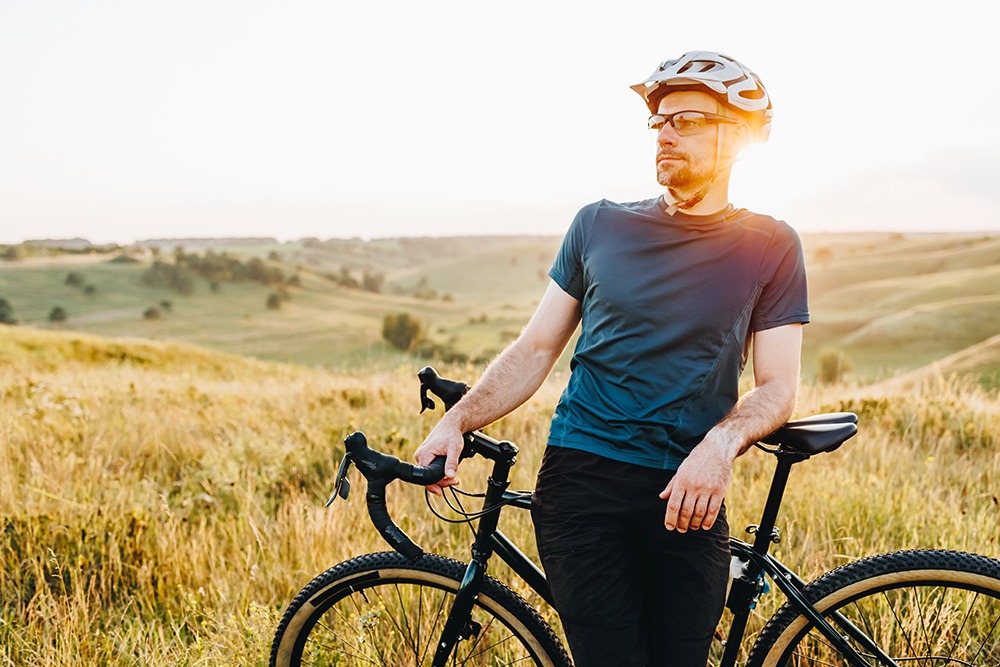 A man wearing a helmet and sunglasses stands with his bicycle in a grassy field at sunset, looking into the distance. Rolling hills and trees are visible in the background.
