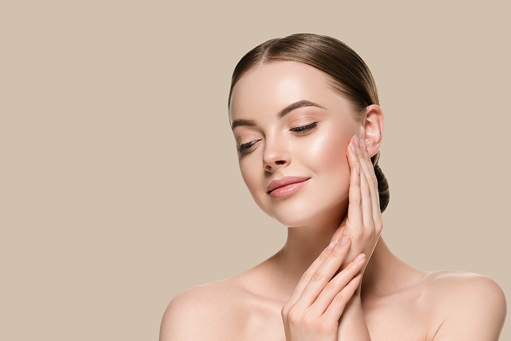 A woman with smooth, glowing skin and brown hair slicked back poses with her eyes closed, gently touching her cheek with both hands against a beige background.