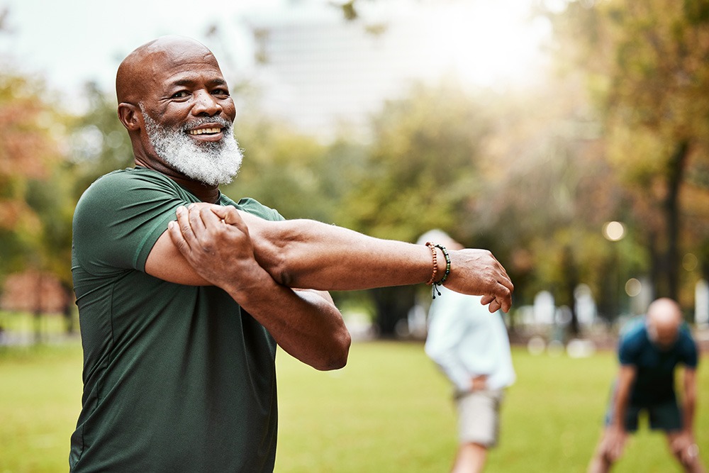 A smiling older man with a gray beard stretches his arm across his chest in a park. He is wearing a green shirt, and other people are stretching in the background among trees.