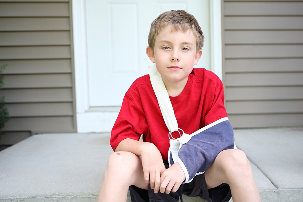 A young boy with short hair sits on a front porch step, wearing a red shirt and black shorts, with his right arm in a blue and white sling. He looks calmly at the camera.