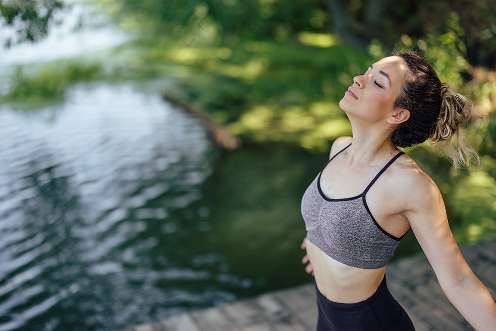A woman in athletic wear stands on a wooden dock by a lake, eyes closed and arms outstretched, enjoying the fresh air and nature around her. Lush green trees and water plants are visible in the background.