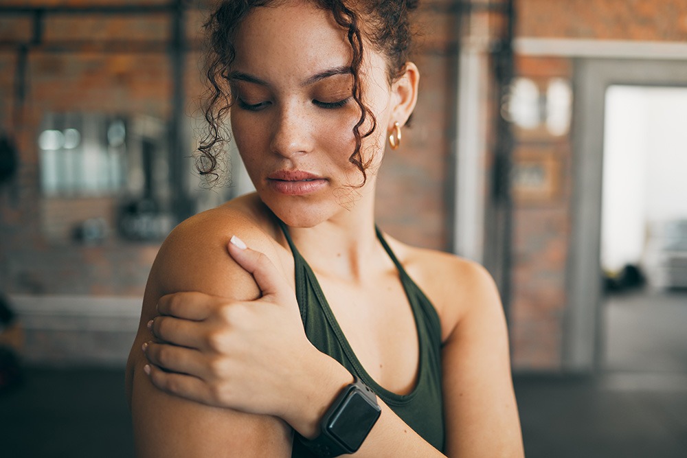A woman in athletic wear stands indoors, gently holding her shoulder with one hand. She has curly hair, is wearing a smartwatch, and appears thoughtful, possibly after a workout. The background is a gym setting.
