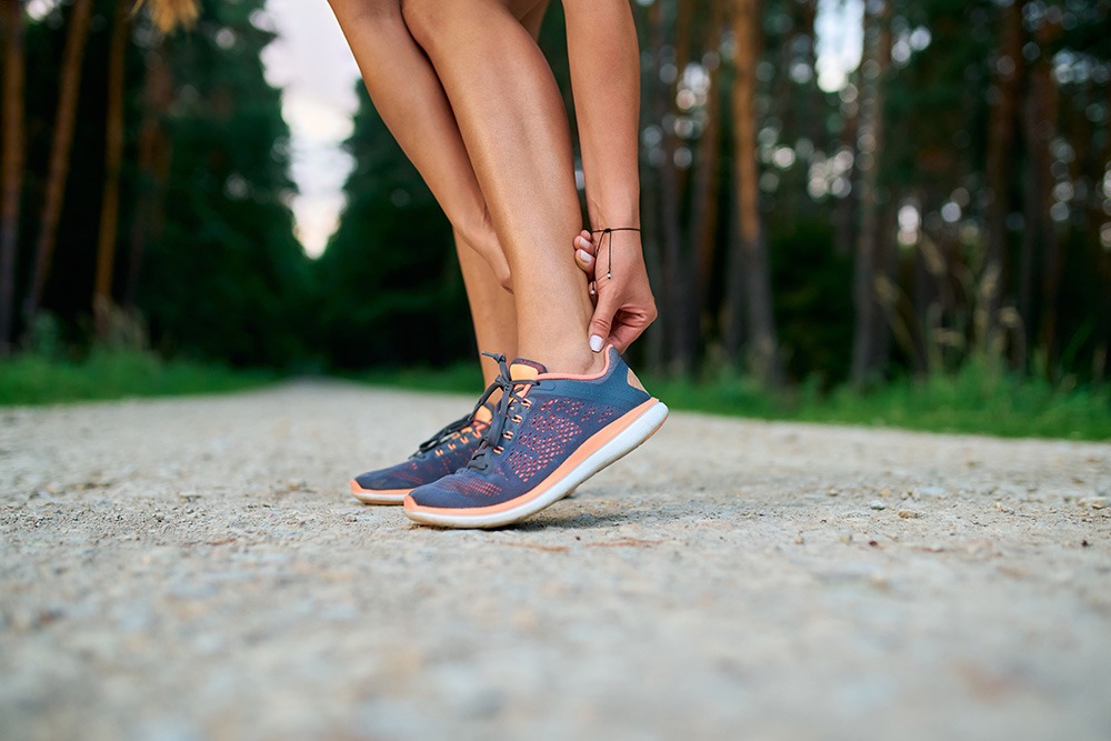 A person standing on a gravel path in a forest, wearing athletic shoes, bends down to touch or check their ankle as if experiencing discomfort or pain.