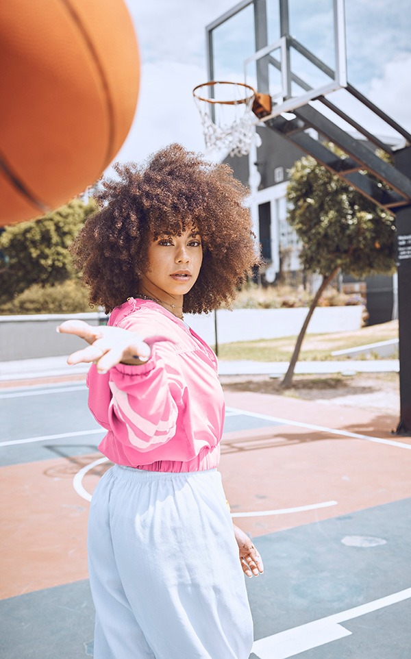 A woman with curly hair wearing a pink jacket and light blue pants is on an outdoor basketball court, tossing a basketball with one hand toward the camera. A basketball hoop is visible in the background.