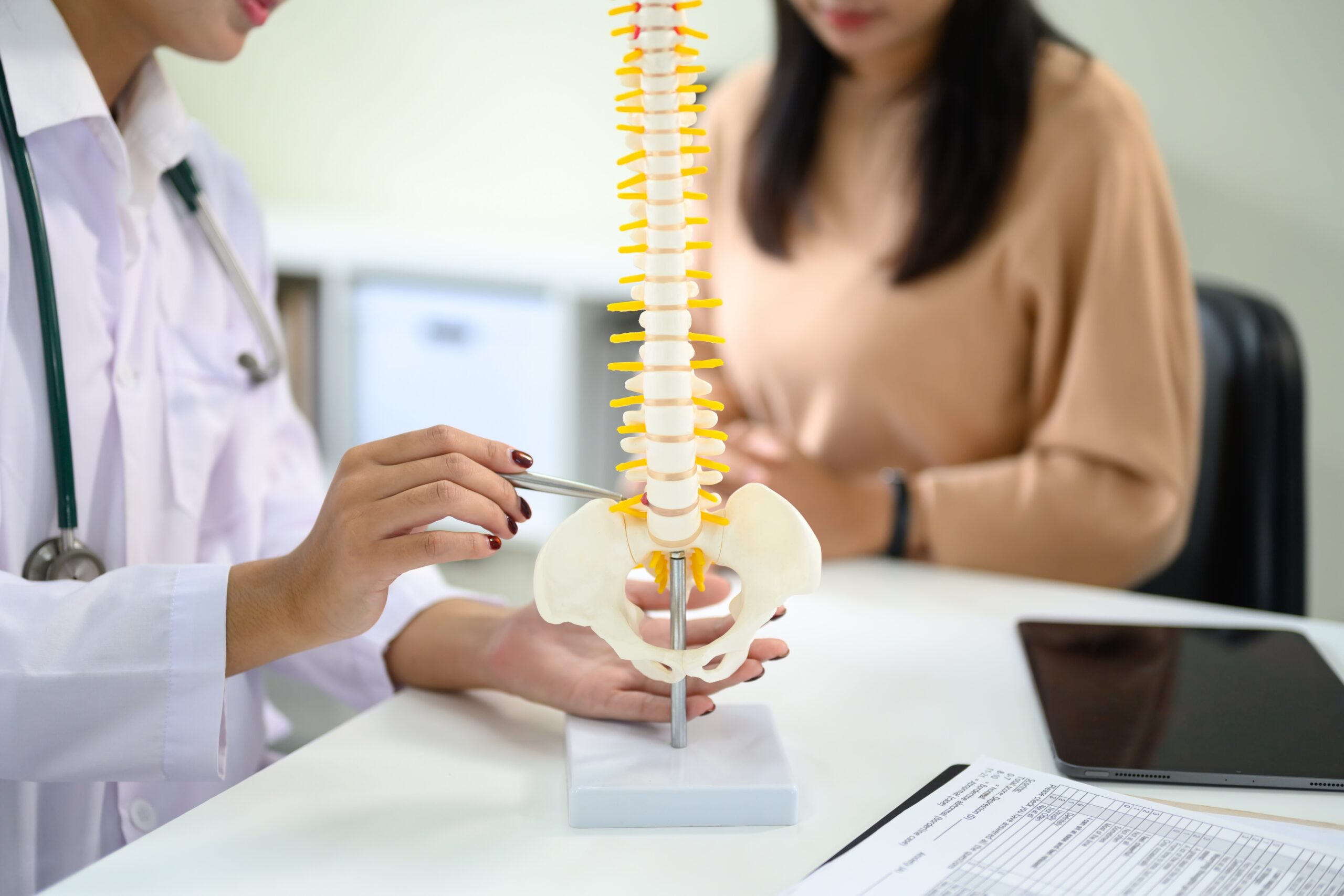A doctor points to a model of the human spine while explaining to a patient. The patient listens attentively, seated across the table. Medical documents and a tablet are on the desk.