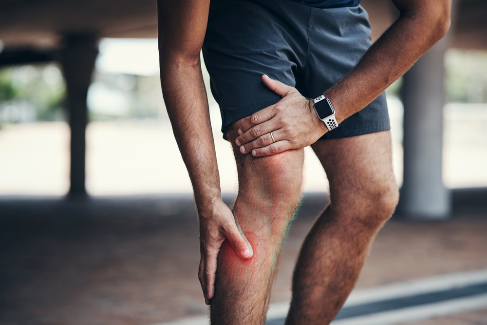 A person in athletic wear holds their knee and shin, highlighting pain and visible veins on the lower leg, suggesting discomfort or injury while exercising outdoors.